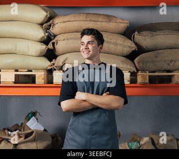 Lagerarbeiter. Lächelnder und selbstbewusster Mann auf einer Schürze, der mit überkreuzten Armen vor Regalen mit Taschen steht. Stockfoto