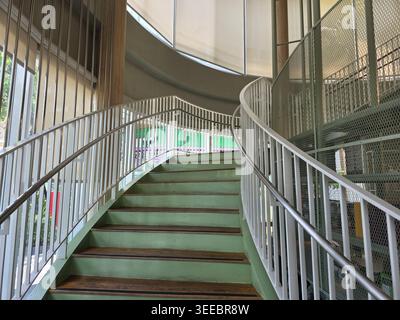 Friedliche geschwungene Treppe mit grüner Treppe und weißem Metallgeländer im modernen Innenraum des Gebäudes. Ruhiger, leerer Raum mit einem einladenden hellen Stockfoto