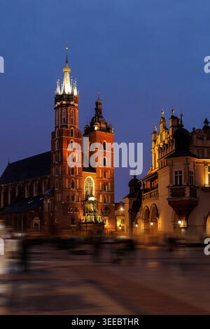 Eine Nachtszene auf dem belebten Krakauer Marktplatz mit bewegungsunscharfen Figuren im Vordergrund. Die beleuchteten Türme der Basilika und das Tuch Ha Stockfoto