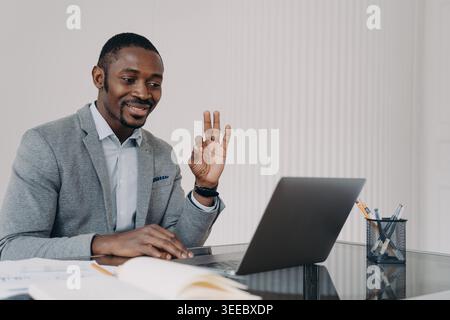 Männlicher afroamerikanischer Manager zeigt OK und genehmigt Geste, mit dem Kunden per Videoanruf am Laptop zu sprechen. Ein lächelnder schwarzer Geschäftsmann zeigt dem Mitarbeiter ein Zeichen für eine Vereinbarung, der online am Computer arbeitet. Stockfoto