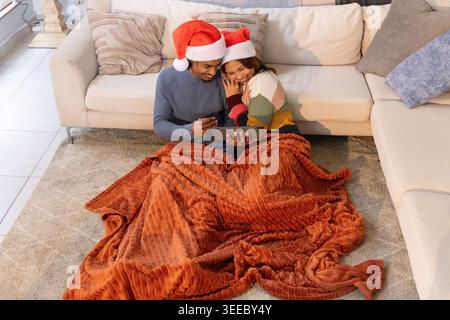 Verschiedene Paare sitzen zu Hause unter Rostdecke und tragen Santa-Hüte, halten Tablette und Karte. Gemütlich, warm, Urlaub, zusammen, intim, Lounge, Kissen Stockfoto