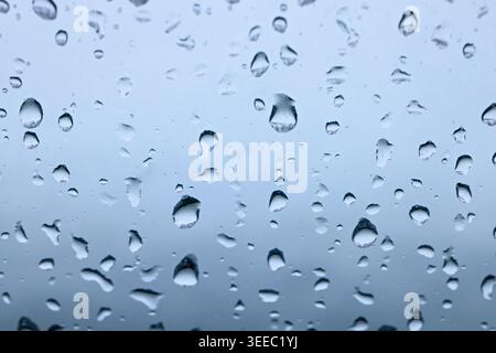 Klare Regentropfen haften vor einem kühlen blauen Hintergrund am Glas und schaffen eine frische, abstrakte Textur für Wetter, Ruhe und saisonale Designthemen. Stockfoto