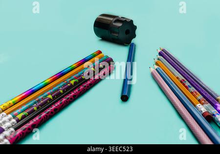 Ein Blick von oben nach unten auf leuchtende, gemusterte Bleistifte, die symmetrisch um einen modernen schwarzen Handspitzer auf einer sauberen türkisfarbenen Oberfläche angeordnet sind. Stockfoto