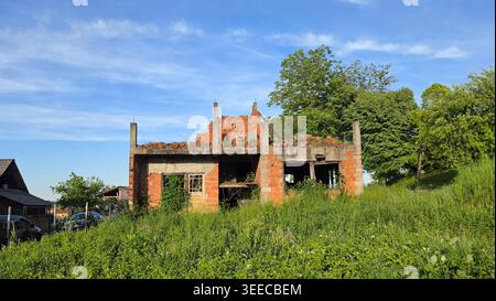 Ein unvollendetes, dachloses Gebäude aus rotem Lehm steht in Vergessenheit inmitten von hohem Unkraut und üppiger grüner Vegetation am Hügel unter einem weichen Nachmittagshimmel Stockfoto