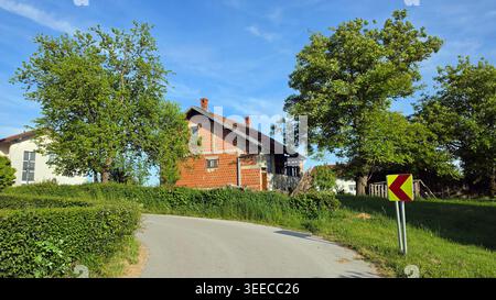 Ein traditionelles Haus aus rotem Lehm befindet sich auf einem grasbewachsenen Hügel, umgeben von grünen Bäumen und einer gepflegten Hecke entlang einer geschwungenen Landstraße mit gelbem Gelb Stockfoto