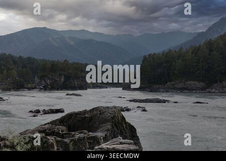 Dramatischer Blick auf den Fluss Katun mit felsigen Stromschnellen, Kiefernwäldern und überlagerten Bergen unter schweren Sturmwolken in der Republik Altai, Russland Stockfoto