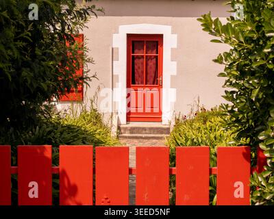 Traditionelles weißes bretonisches Haus mit einer lebendigen roten Holztür, roten Rollläden und rotem Pfahlzaun umgeben von üppiger Gartenvegetation in der Bretagne, Franc Stockfoto
