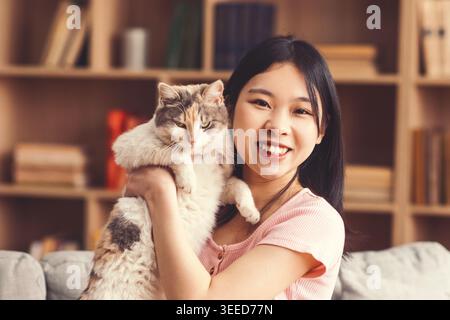 Die junge Frau hält eine Katze und lächelt in einem gemütlichen Innenraum mit Bücherregalen an der Kamera Stockfoto