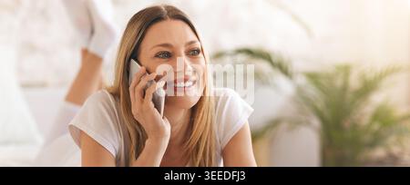 Frau, die auf einem Smartphone spricht, während sie auf einem Bett in einem hellen Raum liegt Stockfoto