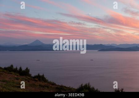 Weite Aussicht auf die Bucht von Avacha bei Sonnenuntergang mit ruhigem Meer, Frachtschiffen und mehrschichtigen Bergsilhouetten unter rosa Wolken, in der Nähe von Petropavlovsk-Kamtschatski, Kamcha Stockfoto