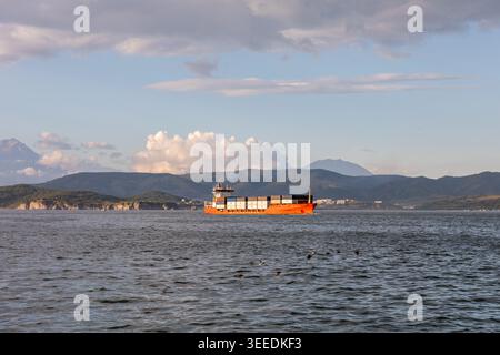 Frachtcontainerschiff auf ruhigem Meer in der Bucht Avacha bei Petropavlovsk-Kamtschatski, Region Kamtschatka, Russland, mit Küstenbergen, Wolken und Abendlicht Stockfoto