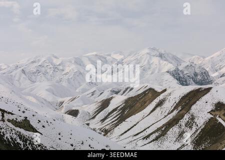 Panoramablick auf schneebedeckte Bergkämme und Gipfel in Kirgisistan, Zentralasien, mit mehrschichtigen Hängen, felsigen Strukturen und einem bewölkten Winterhimmel Stockfoto