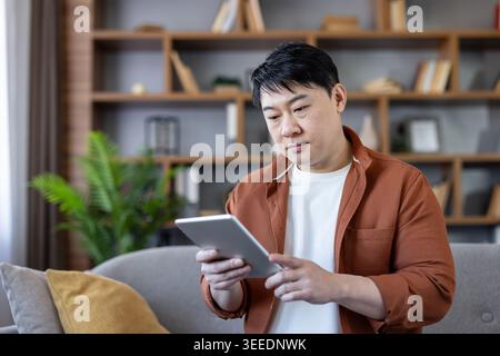 Asiatischer Mann, der zu Hause auf einer bequemen Couch sitzt und ein digitales Tablet für Fernarbeit, Lernen oder Surfen im Internet in einem modernen, gemütlichen Wohnzimmer verwendet Stockfoto