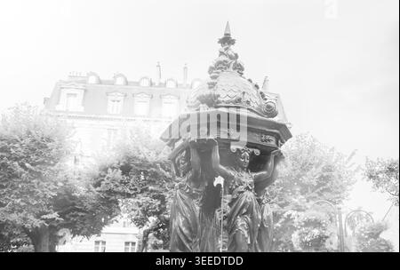 Typischer Pariser Trinkbrunnen mit gusseiserner Frauengruppe auf dem Stadtplatz. Retro-Foto im Alter. Schwarz-weiß. Stockfoto