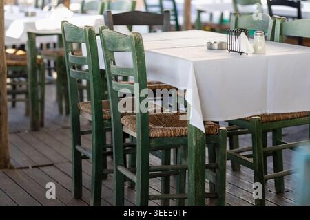 Traditionelle griechische Taverne mit grünen Holzstühlen und gewebten Strohstühlen um einen Tisch mit weißem Tuch auf einer rustikalen Holzterrasse, die einen schafft Stockfoto