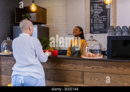 Afroamerikanischer Barista in Senfpullover, der rote Becher auf der Cafettheke mit Gebäckkuppeln serviert Stockfoto