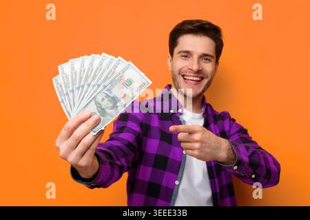 Junge Mann zeigt Cash mit Lächeln vor Orange Hintergrund tagsüber Stockfoto