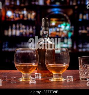 Zwei abgerundete Whiskey-Gläser, Flasche, Eiswürfel und Kristallglas in einer hölzernen Pub-Bar mit dunklen Regalen und warmen Bokeh-Lichtern. Stockfoto