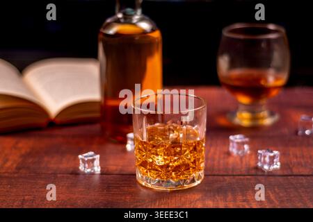 Kristallbecher gefüllt mit Bernstein-Whiskey auf einem hölzernen Pub-Tisch, mit verschwommener Flasche, offenem Buch, Glas und Eiswürfeln in warmem Abendlicht. Stockfoto