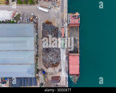 Ein großer Versandcontainer wird an einem Hafen mit Schrott beladen. In der Nähe befinden sich Anhäufungen von Metallschutt und geparkte Fahrzeuge entlang der Küste Stockfoto