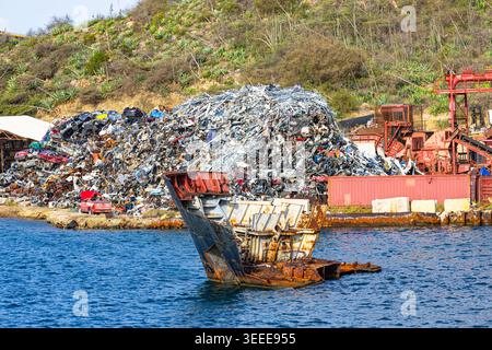 Massiver Haufen von Schrott und zerquetschten Autos an einem Industrieterminal an der Küste mit einem verrosteten Schiffswrack im Wasser Stockfoto