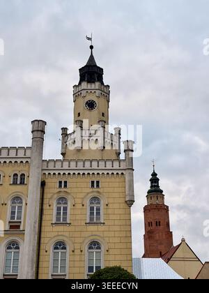 Architektonischer Kontrast zwischen dem neogotischen Rathausuhrturm und dem historischen Backsteinturm der Pfarrkirche in Wschowa, Polen, unter einem bewölkten Himmel. Stockfoto