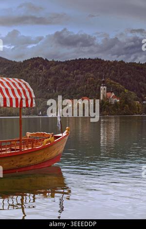 025 modernes Pletna (hölzernes Ruderboot) mit bunt gestreiften Markisen, die mit dem Bug vertäut sind, der auf die Insel in der Mitte des Bleder Sees zeigt. Bled-Slowenien Stockfoto