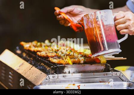 Nahaufnahme: Chefhände, die auf einem heißen Holzkohlegrill Soße auf Fleischspieße gießen. Authentische Street Food Kochhintergründe auf einem Nachtmarkt. Stockfoto