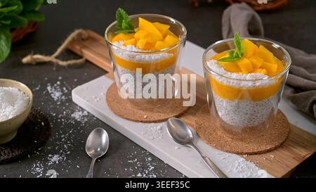 Zwei Gläser Chia Pudding und Mangopüree werden mit frischem Obst und Kokosraspeln garniert. Die rustikale Tischeinstellung verfügt über eine weiche Seitenbeleuchtung Stockfoto
