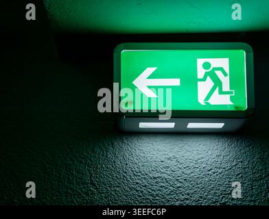 Grün beleuchtetes Notausstiegsschild an einer Wand in einem dunklen Gebäudekorridor. Nahaufnahme eines leuchtenden Notausstiegsschildes. Stockfoto