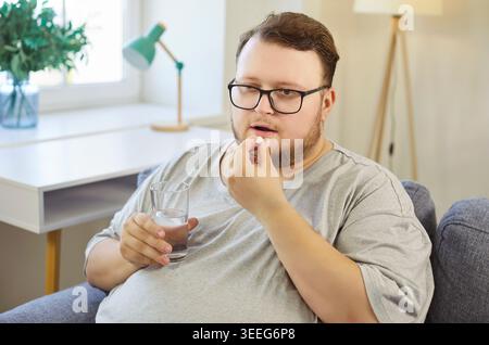 Trauriger übergewichtiger Mann, der Pille zu Hause nimmt, Tablette und Wasserglas in den Händen hält Stockfoto