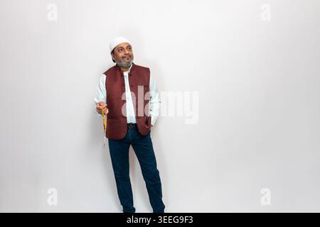 Indischer Mann steht vor einem schlichten Hintergrund, trägt eine traditionelle Weste und Jeans. Er hält einen Rosenkranz in der Hand Stockfoto