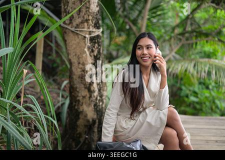 Ein horizontales Medium Shot fängt eine lächelnde Thai-Frau ein, die auf ihrem Smartphone spricht, während sie in einem üppigen Dschungel in Phuket, Thailand, sitzt. ich war angezogen Stockfoto