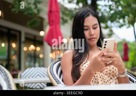 Eine horizontale mittlere Aufnahme erfasst eine wunderschöne Thai-Frau, die sich auf ihr Smartphone konzentriert, während sie sich auf einem Café oder einer Restaurantterrasse in Phuket im Freien entspannt. Stockfoto
