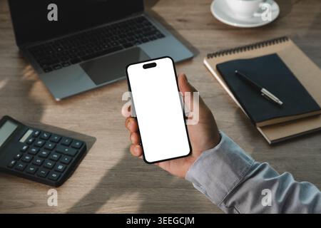 Handheld-Smartphone mit leerem weißen Bildschirm über dem Bürotisch mit Laptop, Rechner und Notebook, für Mockup der mobilen App, Online-Bankin Stockfoto