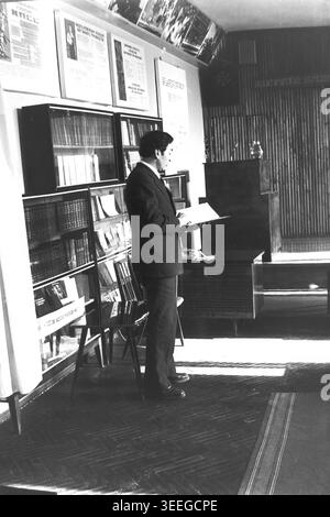 Slaviansk, 1983. Konferenzsaal der Armature-Isolator Factory (AIZ). Ein Mann im Anzug steht an einem großen Bücherregal, liest aus einem Dokument, und die Präsidiumsmitglieder sind im Hintergrund sichtbar. Dieses Vintage-Schwarz-weiß-Foto fängt die authentische Atmosphäre der Epoche ein und spiegelt die täglichen Aktivitäten und Industriekultur der Region während der Sowjetzeit wider. Es dient als wertvolles historisches Dokument der lokalen Gemeinde, das das visuelle Gedächtnis der Menschen und ihres täglichen Lebens bewahrt und einen friedlichen Donbass zeigt. Stockfoto