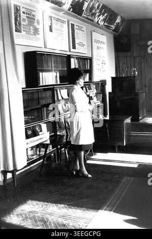 Slaviansk, 1983. Konferenzsaal der Armature-Isolator Factory (AIZ). Eine Frau in einem hellen Mantel steht nachdenklich in der Nähe der Bücherregale während eines offiziellen Bildungs- oder Fabriktreffens. Dieses Vintage-Schwarz-weiß-Foto fängt die authentische Atmosphäre der Epoche ein und spiegelt die täglichen Aktivitäten und Industriekultur der Region während der Sowjetzeit wider. Es dient als wertvolles historisches Dokument der lokalen Gemeinde, das das visuelle Gedächtnis der Menschen und ihres täglichen Lebens bewahrt und einen friedlichen Donbass zeigt. Stockfoto