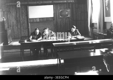 Slaviansk, 1983. Konferenzsaal der Armature-Isolator Factory (AIZ). Ein genauer Blick auf den Präsidiumstisch, wo drei Männer in Anzügen sitzen und Dokumente während eines offiziellen Fabriktreffens überprüfen. Dieses Vintage-Schwarz-weiß-Foto fängt die authentische Atmosphäre der Epoche ein und spiegelt die täglichen Aktivitäten und Industriekultur der Region während der Sowjetzeit wider. Es dient als wertvolles historisches Dokument der lokalen Gemeinde, das das visuelle Gedächtnis der Menschen und ihres täglichen Lebens bewahrt und einen friedlichen Donbass zeigt. Stockfoto