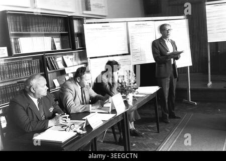 Slaviansk, 1983. Konferenzsaal der Armature-Isolator Factory (AIZ). Ein Redner steht mit Papieren und zeigt auf große Charts auf Ausstellungsständen, die dem sitzenden Präsidium Daten präsentieren. Dieses Vintage-Schwarz-weiß-Foto fängt die authentische Atmosphäre der Epoche ein und spiegelt die täglichen Aktivitäten und Industriekultur der Region während der Sowjetzeit wider. Es dient als wertvolles historisches Dokument der lokalen Gemeinde, das das visuelle Gedächtnis der Menschen und ihres täglichen Lebens bewahrt und einen friedlichen Donbass zeigt. Stockfoto