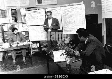 Slaviansk, 1983. Konferenzsaal der Armature-Isolator Factory (AIZ). Ein Redner spricht über Produktionspläne in der Nähe von großen Charts und spricht mit zwei Männern, die mit einem offenen Buch an einem Schreibtisch sitzen. Dieses Vintage-Schwarz-weiß-Foto fängt die authentische Atmosphäre der Epoche ein und spiegelt die täglichen Aktivitäten und Industriekultur der Region während der Sowjetzeit wider. Es dient als wertvolles historisches Dokument der lokalen Gemeinde, das das visuelle Gedächtnis der Menschen und ihres täglichen Lebens bewahrt und einen friedlichen Donbass zeigt. Stockfoto