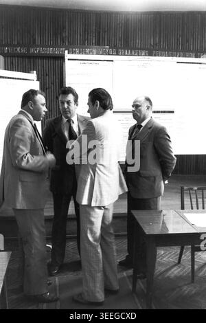 Slaviansk, 1983. Konferenzsaal der Armature-Isolator Factory (AIZ). Vier Männer in formellen Anzügen stehen zusammen und führen ein tiefes Gespräch, wahrscheinlich während einer Pause bei einer offiziellen Veranstaltung. Dieses Vintage-Schwarz-weiß-Foto fängt die authentische Atmosphäre der Epoche ein und spiegelt die täglichen Aktivitäten und Industriekultur der Region während der Sowjetzeit wider. Es dient als wertvolles historisches Dokument der lokalen Gemeinde, das das visuelle Gedächtnis der Menschen und ihres täglichen Lebens bewahrt und einen friedlichen Donbass zeigt. Stockfoto