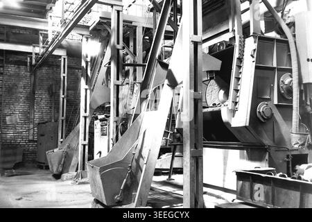 Slaviansk, 1983. In einer schweren Fertigungswerkstatt in der Armature-Isolator Factory (AIZ). Eine massive industrielle Taumelmaschine, die zum Reinigen von Metallgussteilen mit Stahlkugeln verwendet wird, steht im Vordergrund. Es verfügt über tragende Metallbalken und Ladeschaufeln. Dieses Vintage-Schwarz-weiß-Foto fängt die authentische Atmosphäre der Epoche ein und spiegelt die täglichen Aktivitäten und Industriekultur der Region während der Sowjetzeit wider. Es dient als wertvolles historisches Dokument, das einen friedlichen Donbass zeigt. Stockfoto