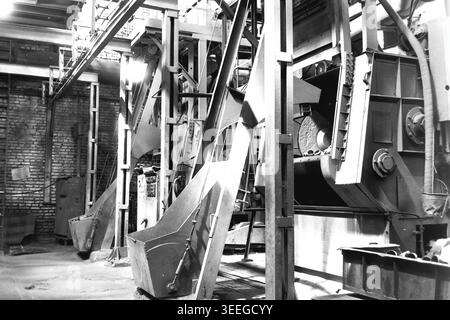 Slaviansk, 1983. In einer schweren Fertigungswerkstatt in der Armature-Isolator Factory (AIZ). Eine massive industrielle Taumelmaschine, die zum Reinigen von Metallgussteilen mit Stahlkugeln verwendet wird, steht im Vordergrund. Es verfügt über tragende Metallbalken und Ladeschaufeln. Dieses Vintage-Schwarz-weiß-Foto fängt die authentische Atmosphäre der Epoche ein und spiegelt die täglichen Aktivitäten und Industriekultur der Region während der Sowjetzeit wider. Es dient als wertvolles historisches Dokument, das einen friedlichen Donbass zeigt. Stockfoto