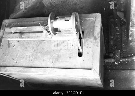 Slaviansk, 1983. Arbeitsstation in der Armature-Insulator Factory (AIZ). Nahaufnahme einer kleinen, speziell angefertigten mechanischen Vorrichtung oder Drehmaschine mit Riemenantrieb und angeflanschtem Rad, sicher auf einer Werkbank aus Schwermetall montiert. Dieses Vintage-Schwarz-weiß-Foto fängt die authentische Atmosphäre der Epoche ein und spiegelt die täglichen Aktivitäten und Industriekultur der Region während der Sowjetzeit wider. Es dient als wertvolles historisches Dokument der lokalen Gemeinde, bewahrt das visuelle Gedächtnis und zeigt einen friedlichen Donbass. Stockfoto