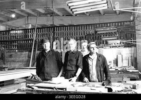 Slaviansk, 1983. In einer Werkstatt im Armature-Isolator Plant (AIZ). Männliche Handwerker stehen selbstbewusst hinter einer Werkbank, die mit Holzteilen und Werkzeugen bedeckt ist, und posieren für ein Porträt im gut beleuchteten Arbeitsbereich. Dieses Vintage-Schwarz-weiß-Foto fängt die authentische Atmosphäre der Epoche ein und spiegelt die täglichen Aktivitäten und Industriekultur der Region während der Sowjetzeit wider. Es dient als wertvolles historisches Dokument der lokalen Gemeinde, bewahrt das visuelle Gedächtnis der Menschen und zeigt einen friedlichen Donbass. Stockfoto