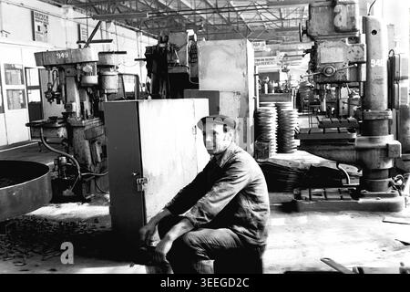 Slaviansk, 1983. In einer Bearbeitungswerkstatt im Armature-Isolator Plant (AIZ). Ein männlicher Arbeiter sitzt zwischen schweren Bohrpressen und verstreuten Metallteilen und macht eine kurze Pause in der geschäftigen Fabrikhalle. Dieses Vintage-Schwarz-weiß-Foto fängt die authentische Atmosphäre der Epoche ein und spiegelt die täglichen Aktivitäten und Industriekultur der Region während der Sowjetzeit wider. Es dient als wertvolles historisches Dokument der lokalen Gemeinde, bewahrt das visuelle Gedächtnis der Menschen und zeigt einen friedlichen Donbass. Stockfoto
