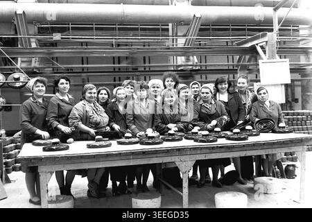 Slaviansk, 1983. In der Armature-Isolator Plant (AIZ). Ein großes Team lächelnder Arbeiterinnen posiert zusammen für ein Gruppenporträt hinter einem langen Holztisch, gefüllt mit großen, dunklen keramischen industriellen elektrischen Isolatoren. Dieses Vintage-Schwarz-weiß-Foto fängt die authentische Atmosphäre der Epoche ein und spiegelt die täglichen Aktivitäten und Industriekultur der Region während der Sowjetzeit wider. Es dient als wertvolles historisches Dokument der lokalen Gemeinde, bewahrt das visuelle Gedächtnis der Menschen und zeigt einen friedlichen Donbass. Stockfoto