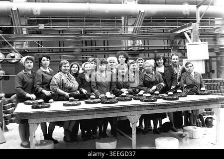 Slaviansk, 1983. In der Armature-Isolator Plant (AIZ). Ein großes Team lächelnder Arbeiterinnen posiert zusammen für ein Gruppenporträt hinter einem langen Holztisch, gefüllt mit großen, dunklen keramischen industriellen elektrischen Isolatoren. Dieses Vintage-Schwarz-weiß-Foto fängt die authentische Atmosphäre der Epoche ein und spiegelt die täglichen Aktivitäten und Industriekultur der Region während der Sowjetzeit wider. Es dient als wertvolles historisches Dokument der lokalen Gemeinde, bewahrt das visuelle Gedächtnis der Menschen und zeigt einen friedlichen Donbass. Stockfoto