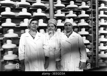 Slaviansk, 1983. In der Armature-Isolator Plant (AIZ). Drei weibliche Arbeiter in weißen Labormänteln und Kopftüchern posieren selbstbewusst für ein Porträt vor hohen Racks, die mit ungebrannten weißen glockenförmigen Isolatoren aus Keramik gefüllt sind. Dieses Vintage-Schwarz-weiß-Foto fängt die authentische Atmosphäre der Epoche ein und spiegelt die täglichen Aktivitäten und Industriekultur der Region während der Sowjetzeit wider. Es dient als wertvolles historisches Dokument der lokalen Gemeinde, bewahrt das visuelle Gedächtnis und zeigt einen friedlichen Donbass. Stockfoto