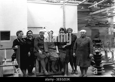 Slaviansk, 1983. In der Armature-Isolator Plant (AIZ). Eine freundliche Gruppe weiblicher Arbeiter sitzt und steht um einen schweren Industriecontainer voller dunkler Keramikisolatoren und lächelt warm für die Kamera. Dieses Vintage-Schwarz-weiß-Foto fängt die authentische Atmosphäre der Epoche ein und spiegelt die täglichen Aktivitäten und Industriekultur der Region während der Sowjetzeit wider. Es dient als wertvolles historisches Dokument der lokalen Gemeinde, bewahrt das visuelle Gedächtnis und zeigt einen friedlichen Donbass. Stockfoto